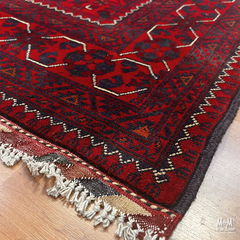 A close-up of a red patterned rug with dark navy and beige geometric motifs, shown on a light wood floor with fringed tassels visible at one corner.